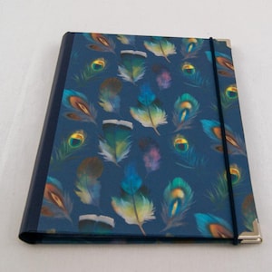 May include: A rectangular notebook with a dark blue cover featuring a repeating pattern of colorful peacock feathers in shades of teal, green, and gold. The notebook has a solid dark blue spine, silver-toned metal corner protectors, and a black elastic band.