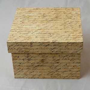Peut inclure: Une boîte de rangement carrée avec couvercle, présentant une surface de couleur crème recouverte d'un motif répétitif d'écriture cursive noire rappelant une écriture historique, avec des tons de papier vieilli brun sur tout l'extérieur.