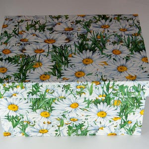 Può includere: Una scatola rettangolare con coperchio, decorata con un motivo di margherite bianche con centri gialli e foglie verdi. La scatola è posta su uno sfondo bianco, evidenziando il design floreale.