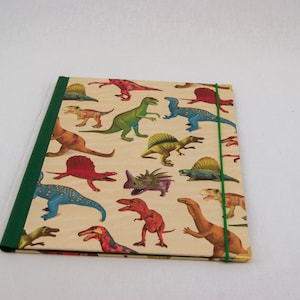 May include: A rectangular folder with a beige background featuring a repeating pattern of various colorful dinosaurs in shades of red, green, and blue. The folder has a solid dark green spine and a thin green elastic band closure on the right side.