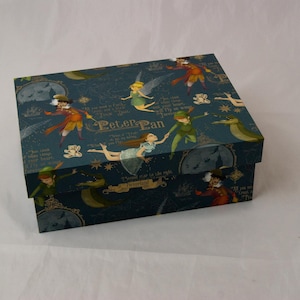 May include: A rectangular storage box with a dark blue background featuring a Peter Pan theme. The lid and sides display illustrations of Peter Pan, Wendy, Tinker Bell, and Captain Hook, along with the text Peter Pan and Neverland.