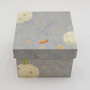 May include: A square, light blue storage box featuring illustrations from The Little Prince, including yellow stars, planets, and the character holding balloons. The text Le Petit Prince is printed on the surface in a cursive, dark gray font.