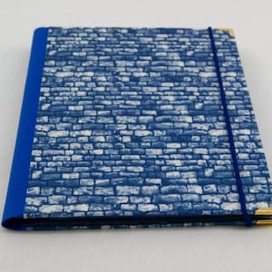 Puede incluir: Un cuaderno con estampado azul y blanco, cierre elástico azul y esquinas de metal dorado.