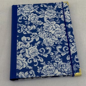 May include: A rectangular notebook featuring a deep blue cover with a white floral pattern. The spine is solid blue, and the cover is secured by a vertical blue elastic band. The corners are reinforced with gold-toned metal accents.