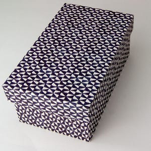 Puede incluir: Caja de regalo rectangular con tapa. La caja está cubierta con un patrón repetitivo de triángulos blancos y azul marino. La caja está sobre una superficie blanca.