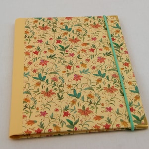 May include: A rectangular notebook with a cream-colored cover featuring a repeating floral pattern in shades of red, orange, and green. The notebook has a solid yellow spine, gold-toned metal corner protectors, and a thin, light blue elastic band.