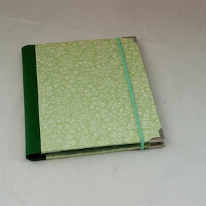 May include: A rectangular notebook featuring a light green cover with a white floral pattern and a solid dark green spine. The notebook is secured with a light green elastic band and includes silver-toned metal corner protectors on the front cover.
