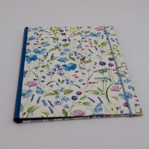 Könnte beinhalten: Eine rechteckige Mappe mit weißem Einband, der mit einem bunten Aquarellmuster aus blauen, violetten und rosafarbenen Wildblumen sowie grünem Blattwerk bedruckt ist. Die Mappe hat einen dunkelblauen Buchrücken und ein blaues Gummiband.