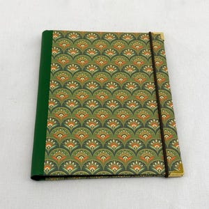 May include: A rectangular notebook featuring a green spine and a cover with a repeating fan-shaped pattern in shades of green, orange, and white. A thin dark brown elastic band wraps vertically around the cover, which has gold-toned metal corners.