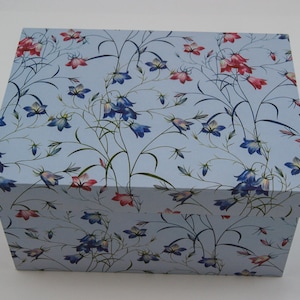 Könnte beinhalten: Eine rechteckige Aufbewahrungsbox mit hellblauem Hintergrund und einem wiederkehrenden Muster aus zarten, glockenförmigen blauen und rosa Blüten mit dünnen grünen Stielen und Blättern. Die Box verfügt über einen passenden Deckel, der den oberen Teil bedeckt.