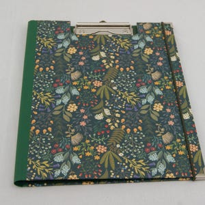 Könnte beinhalten: Ein Klemmbrett mit dunkelblau-grüner floraler Musterabdeckung mit einer Vielzahl von Blumen und Blättern in Gelb-, Rosa- und Blautönen. Der Buchrücken ist grün, und eine silberne Klammer hält die Seiten. Ein Gummiband sichert das Klemmbrett.