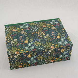 May include: A square box with a dark green background and a floral pattern featuring yellow, blue, and red flowers, and green leaves. The box has a green border around the top edge.