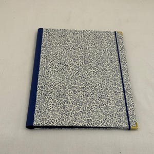 May include: A rectangular portfolio folder featuring a white cover with a dense navy blue floral pattern. The spine is solid navy blue, and the folder is secured by a thin navy blue elastic band. Gold-toned metal corners protect the edges.
