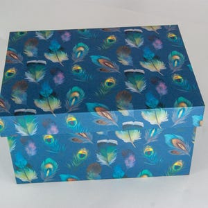 May include: A rectangular storage box with a lid, featuring a dark blue background patterned with a repeating design of colourful peacock feathers in shades of teal, green, gold, and purple, covering both the top lid and the sides of the box.