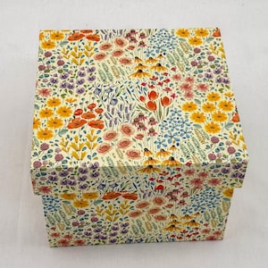 Pode incluir: Uma caixa de armazenamento quadrada com um fundo creme e uma estampa botânica densa e colorida. O padrão inclui ilustrações em estilo aquarela de papoulas laranja, margaridas amarelas, rosas cor-de-rosa, lavanda roxa e flores silvestres azuis.