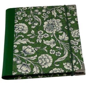May include: A rectangular notebook featuring a green spine and a cover with a white floral pattern on a green background. The notebook is secured with a thin green elastic band and includes silver-toned metal corner protectors on the right side.