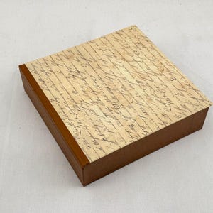 Puede incluir: Una caja cuadrada marrón con una tapa con un diseño de escritura vintage. La caja tiene una base marrón y una tapa con un fondo color crema cubierto con escritura negra descolorida. La caja es para almacenamiento o exhibición.