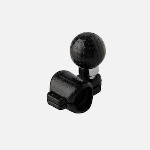 May include: A black steering wheel spinner knob with a textured grip. The knob is round and has a dimpled surface, mounted on a clamp-style base. The base is also black and has a textured surface.