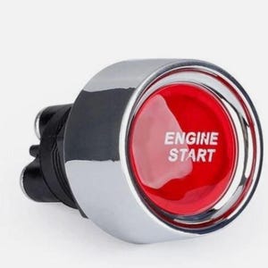 May include: A shiny chrome engine start button with a vibrant red centre. The centre of the button features the words "ENGINE START" in white text. The button has a black base with three prongs.