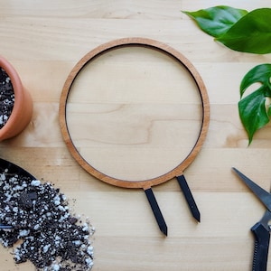 May include: A wooden plant hanger with two black metal stakes. The hanger is round and has a natural wood finish. The stakes are used to secure the hanger to a pot.
