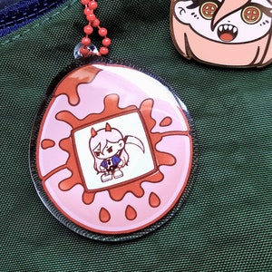 Puede incluir: Un amuleto de acrílico rosa y rojo con un personaje de dibujos animados en su interior. El amuleto está unido a una cadena roja. También se muestra un pin de esmalte rosa y dorado separado con un personaje de dibujos animados.