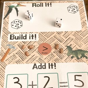 Puede incluir: Una pizarra blanca con las palabras "Roll It!" y "Build It!" escritas en negro. Hay dos dados con puntos negros en ellos. La pizarra también tiene las palabras "Add It!" y un problema de matemáticas escrito en negro: 3 + 2 = 5. También hay imágenes de un dinosaurio y un martillo.