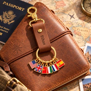 May include: A brown leather keychain with a gold-tone metal lobster clasp and ring. The leather strap is engraved with the name Ethan. Attached to the gold ring are six small square charms featuring the flags of the USA, UK, China, Italy, France, and Spain.
