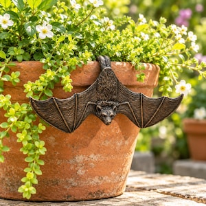 May include: A bronze-colored bat figurine with outstretched wings, designed to hang over the rim of a terracotta flower pot. The bat features detailed fur texture on its body and defined wing membranes, positioned against a backdrop of green foliage.