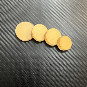 May include: Four round, light brown wooden discs of varying sizes are arranged in a row. The discs are made of a light-coloured wood and have a smooth surface. The background is a black, textured surface.