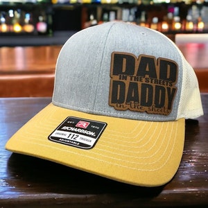 Può includere: Un cappello da camionista grigio e beige con una toppa in pelle marrone che dice "Dad in the streets Daddy in the sheets". Il cappello ha una visiera gialla e un cinturino regolabile nero con il logo Richardson e il testo "112 Trucker".
