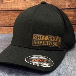May include: Black baseball cap with a curved peak. The cap has a brown rectangular patch with the text "SHIT SHOW SUPERVISOR". A Flexfit label is on the peak. The cap is a Flexfit S/M size.