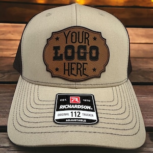 Puede incluir: Una gorra de camionero beige y negra con un parche de cuero marrón que dice "YOUR LOGO HERE" en letras negras. La gorra es ajustable y tiene un logotipo de Richardson en la parte posterior.