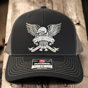 以下が含まれることがあります： 黒と灰色のトラッカーハットに、銀色の鷲のグラフィックと「We the People Shall Not Be Infringed」の文字がプリントされています。帽子は調節可能で、リチャードソンのロゴが付いています。
