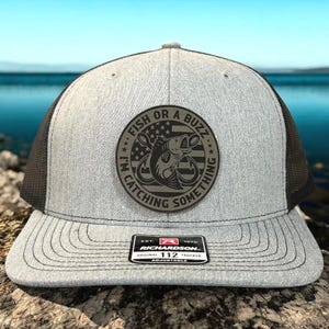 May include: Gray trucker hat with a black mesh back and a curved brim. The front features a circular patch with a fish graphic and the text "FISH OR A BUZZ... I'M CATCHING SOMETHING." A small rectangular patch with text is on the brim.