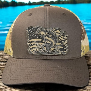 May include: A brown trucker hat with a patch featuring a leaping fish in front of a distressed American flag. The hat has a mesh back with a camouflage pattern and a curved brim. The hat is sitting on a wooden surface.