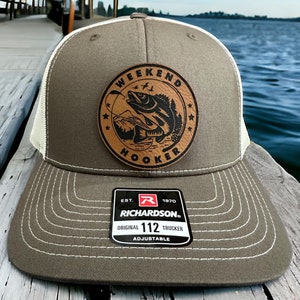 May include: A brown and white trucker hat with a brown leather patch that says "Weekend Hooker" with a fish graphic. The hat is made by Richardson and is adjustable.