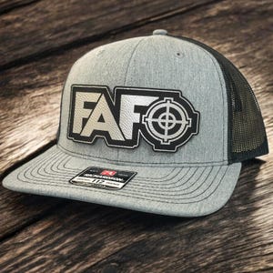 Op de afbeelding: Een grijze en zwarte trucker-pet met een zilveren en zwarte patch waarop "FAFO" staat met een doelwitsymbool.