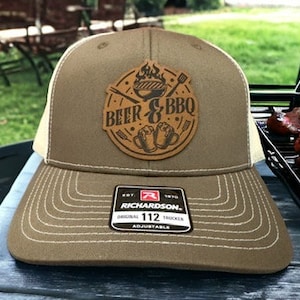 Op de afbeelding: Een bruine en beige trucker-pet met een bruin leren patch waarop "Beer & BBQ" staat met een grill- en vlammenontwerp. De pet is verstelbaar en heeft een mesh-rug.