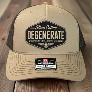 Könnte beinhalten: Beige und schwarze Trucker-Mütze mit dem Text "Blue Collar DEGENERATE" und den Sätzen "STILL WORKING | STILL DIRTY | STILL BROKE". Die Mütze hat ein Richardson 112 Trucker-Etikett.
