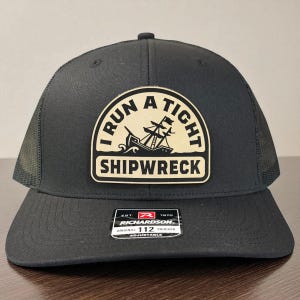 May include: Dark grey trucker cap with a curved peak and mesh back. The cap has a patch with the text "I RUN A TIGHT SHIPWRECK" and a ship graphic. The Richardson brand label is visible.