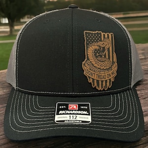 May include: A black and gray trucker hat with a brown leather patch featuring a coiled snake and an American flag with the text "Don't Tread On Me". The hat has a black and gray mesh back and an adjustable strap with the Richardson 112 logo.