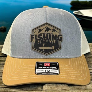 Peut inclure: Casquette de camionneur avec une couronne grise, une visière beige et un dos en filet blanc. Le chapeau présente un écusson avec le texte "I'D RATHER BE FISHING" et un graphique de montagnes, d'arbres et d'un bateau. Le chapeau a une étiquette Richardson 112.