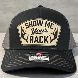 Può includere: Cappellino da camionista nero e grigio con la scritta "SHOW ME Your RACK" su una toppa beige con grafica di corna. Il cappellino ha una visiera curva e una vestibilità regolabile.