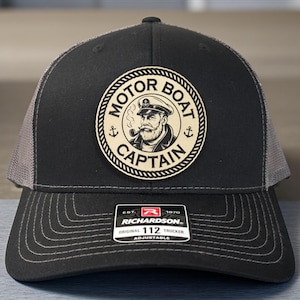May include: Black and gray trucker hat with a patch that reads "MOTOR BOAT CAPTAIN" around an illustration of a captain smoking a pipe. The hat has a curved brim and a mesh back. The hat is adjustable.