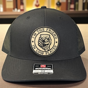 May include: Dark grey trucker cap with a mesh back and a curved peak. The cap has a circular patch with the text "I'M THE FRIEND IN LOW PLACES" and a graphic of a hand holding a glass of whiskey. It is adjustable.