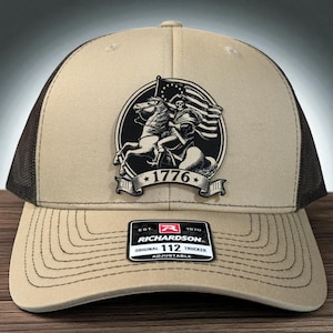 May include: Tan and brown trucker hat featuring a black and white graphic of a skeleton on horseback with a flag. The hat displays the text "1776" and a Richardson 112 Trucker patch.