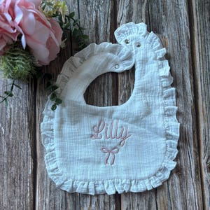 May include: White baby bib with a frilled edge and popper fastening. The bib has the name "Lilly" embroidered in pink script with a bow design. Made from a soft, textured fabric.
