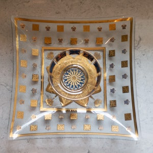 Puede incluir: Plato cuadrado de vidrio con borde dorado y diseño central decorativo dorado y azul. El plato presenta un patrón repetitivo de cuadrados dorados y formas de corona en los bordes. El centro tiene un diseño detallado y ornamentado.
