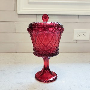 May include: A ruby red, lidded candy dish with a pedestal base. The glass dish features ornate, embossed detailing, including floral and geometric patterns. The lid has a small, round finial. The dish is displayed on a white countertop.
