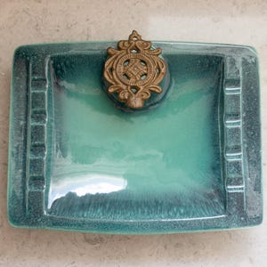 May include: A rectangular ashtray with a teal and turquoise glaze. The ashtray has four cigarette rests and a decorative brass emblem. The ashtray is a vintage item.